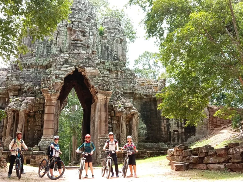 Five Social Cycles riders with their bikes in front of a carved stone face-tower gate at Angkor