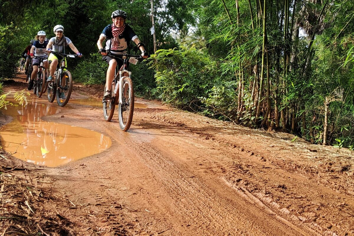 Riders coming along a red dirt track lined with bamboo, past a large puddle