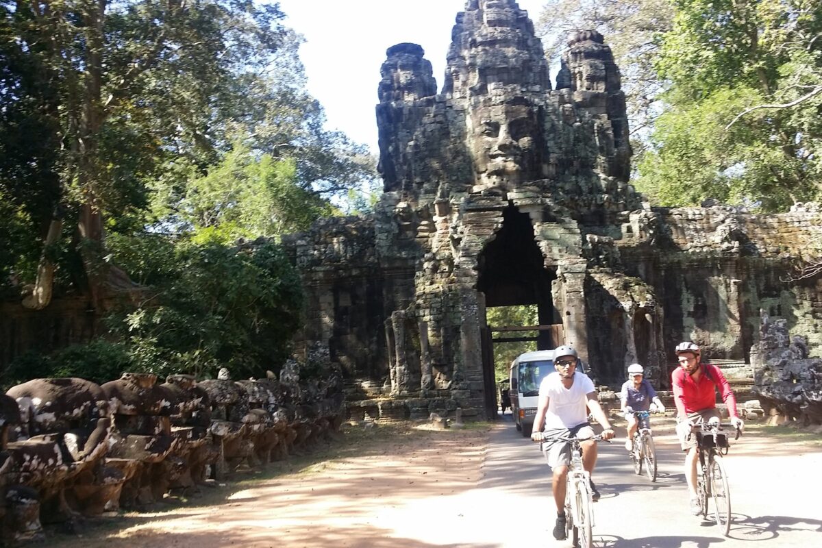 Riders cycling out of an Angkor Thom face-tower gate past the row of stone figures