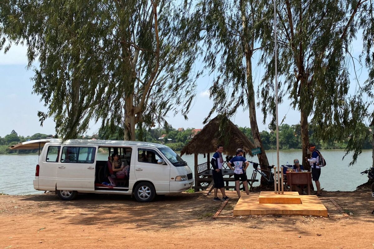 The support van parked under tall trees beside a wide river while riders take a break