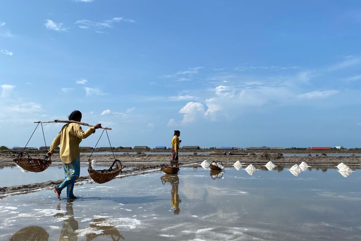 Salt workers carrying baskets on a shoulder pole across flooded salt pans under a blue sky