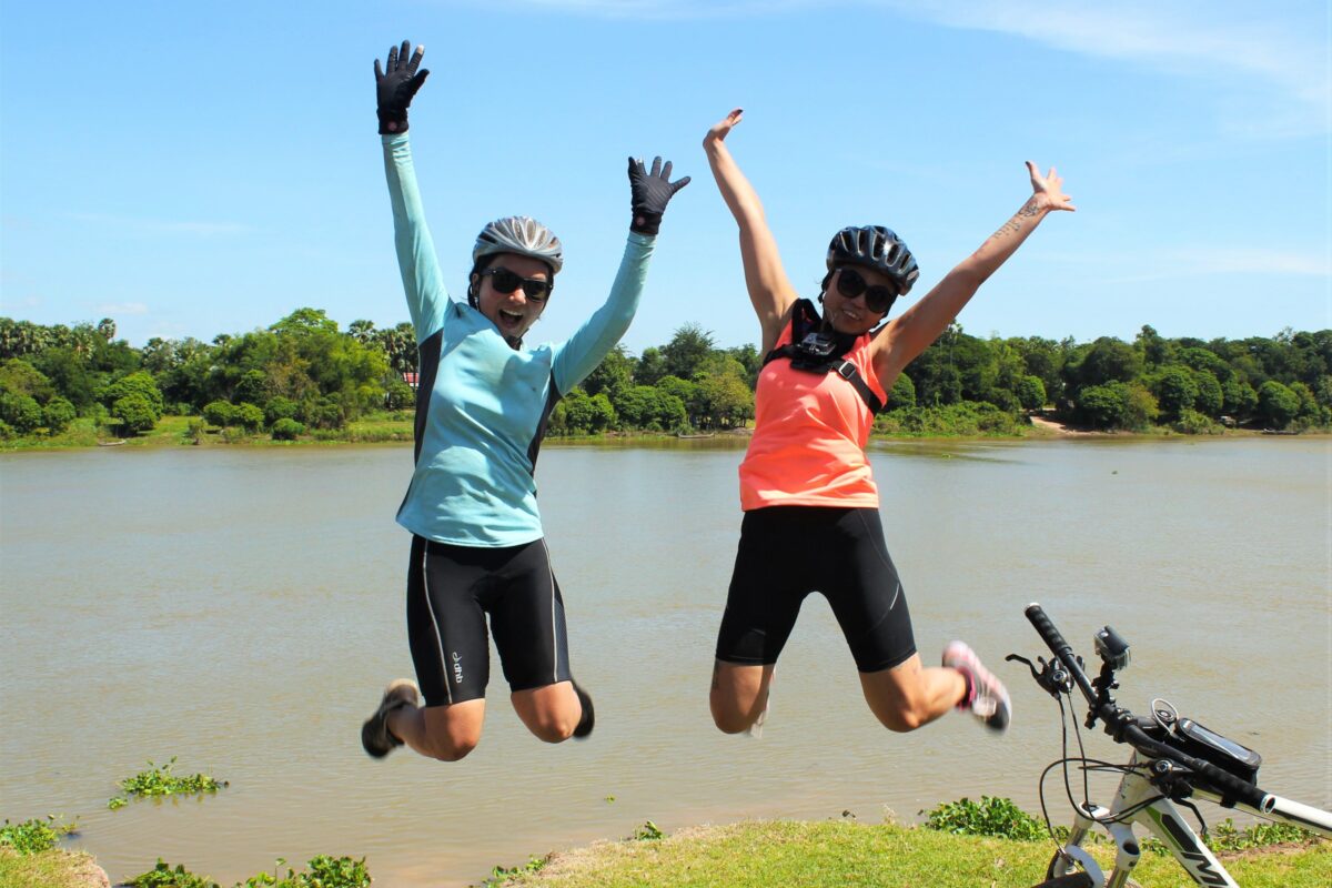 Two riders jumping for joy on a grassy riverbank, a bike in the foreground