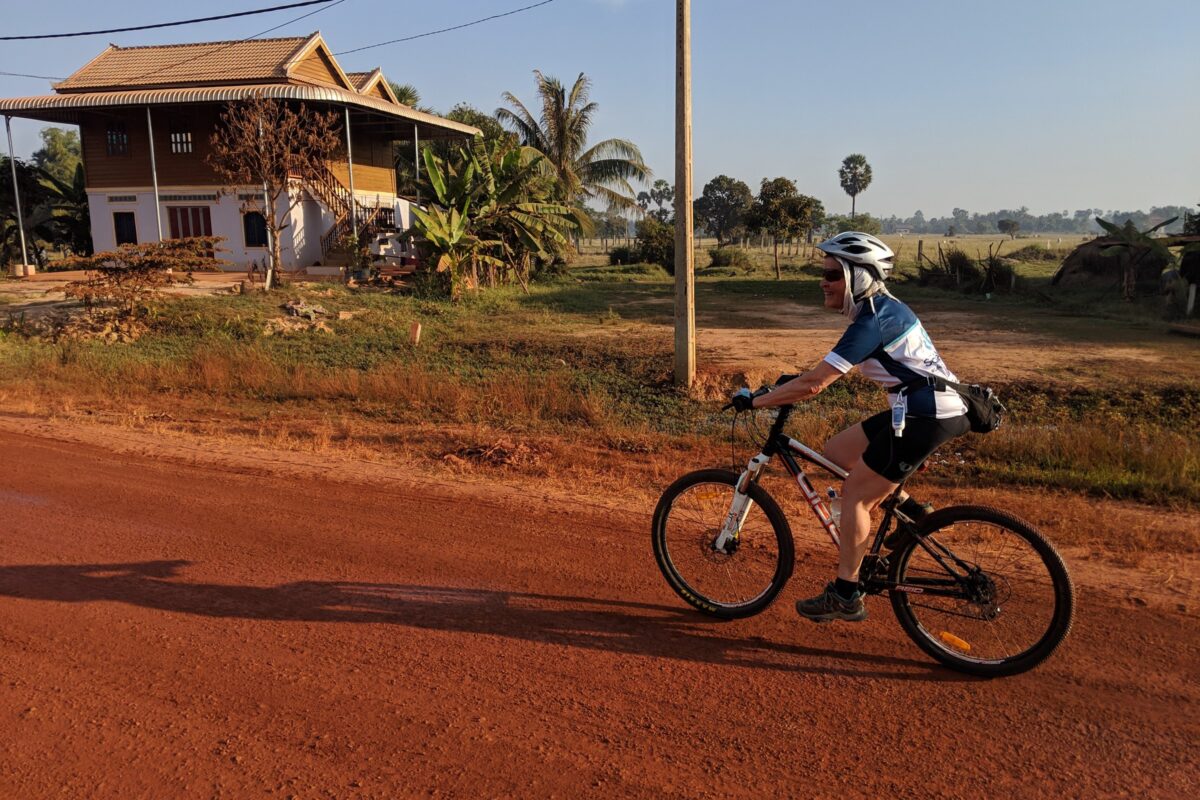 A rider passes a stilt house on a red laterite road in low sun