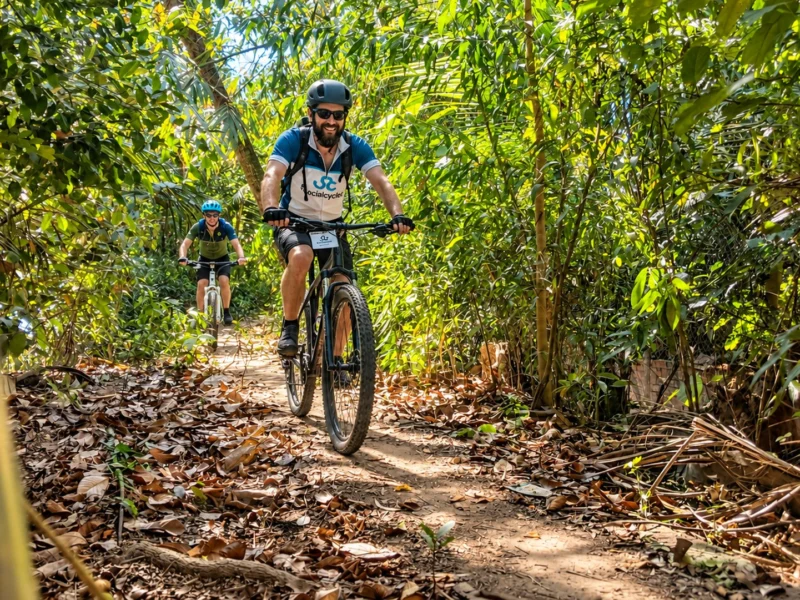 Two riders on a leafy dirt track between palms and bamboo, the lead rider in a Social Cycles jersey smiling at the camera