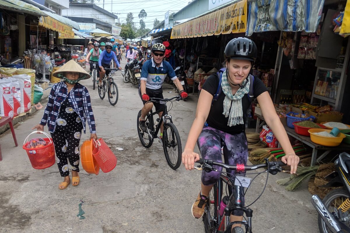 Riders pass a woman carrying baskets through a village market street