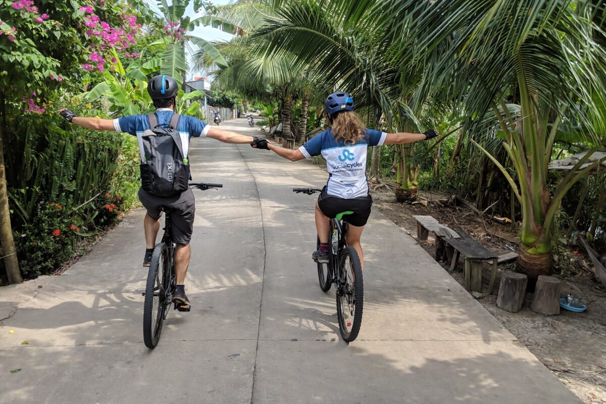 Two riders, arms out, on a lane under coconut palms and bougainvillea