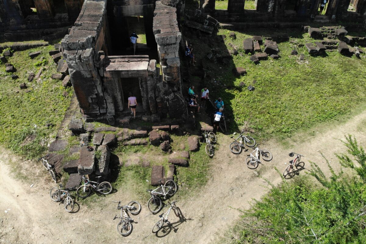 From the air: riders explore a ruined temple, bikes laid in the grass