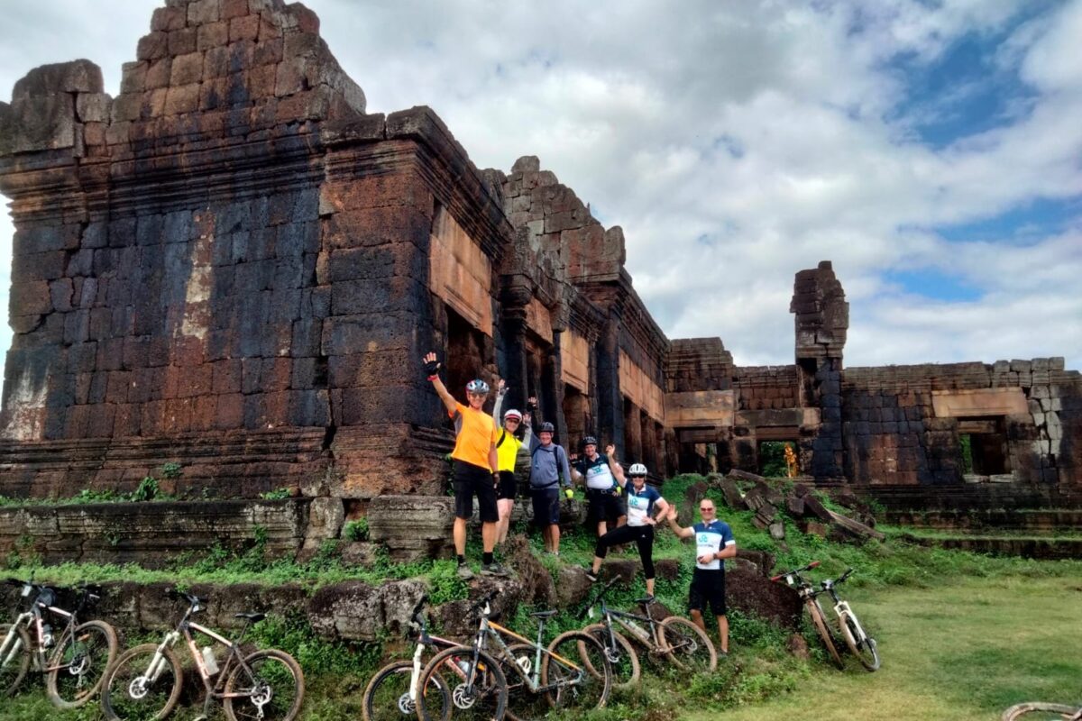 Arms up in front of a towering laterite temple ruin, bikes in the grass