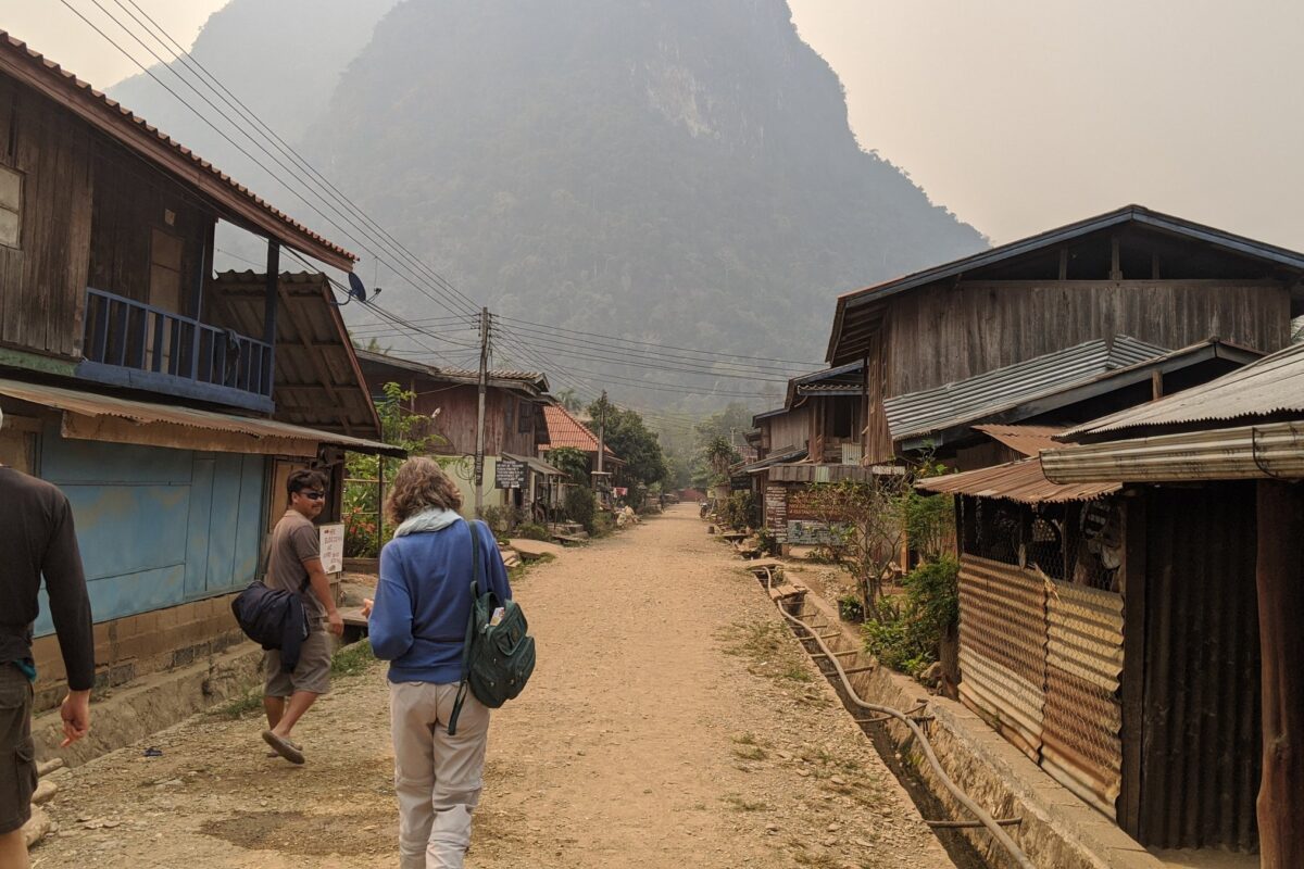 Travellers on a dirt village street beneath a karst peak, Muang Ngoi