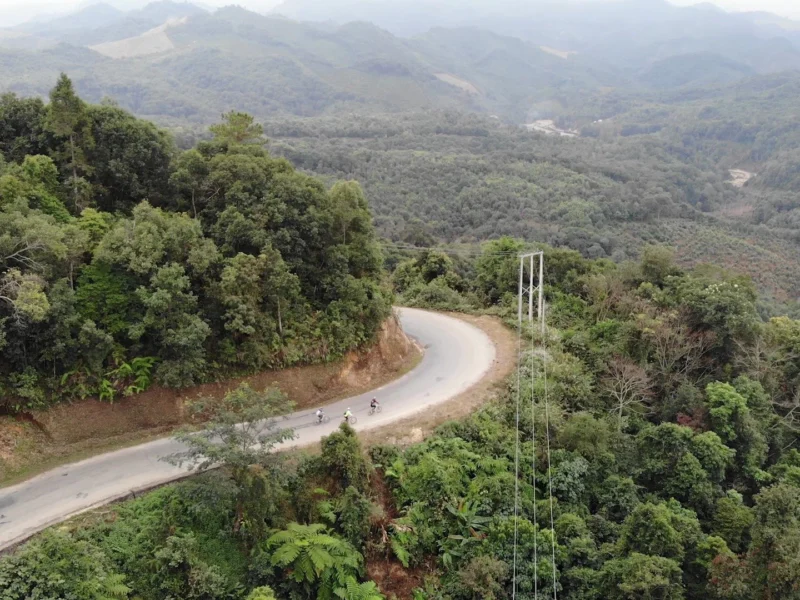From the air: riders small on a hairpin road through forested hills