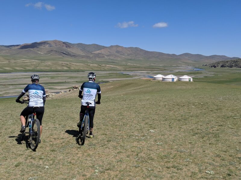 Two riders in Social Cycles jerseys, seen from behind, ride across open grassland towards a river valley with a small ger camp and hills beyond