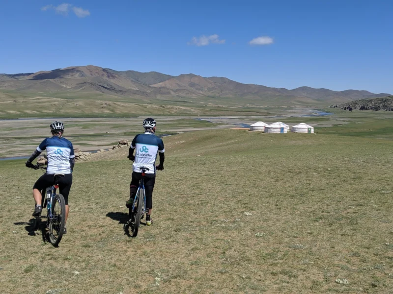 Two riders in Social Cycles jerseys, seen from behind, ride across open grassland towards a river valley with a small ger camp and hills beyond