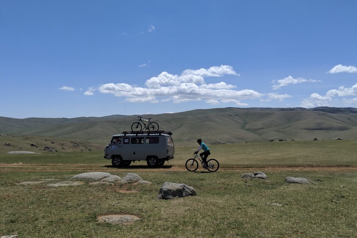 The support van, a bike on its roof, rolls beside a rider in a wide green valley